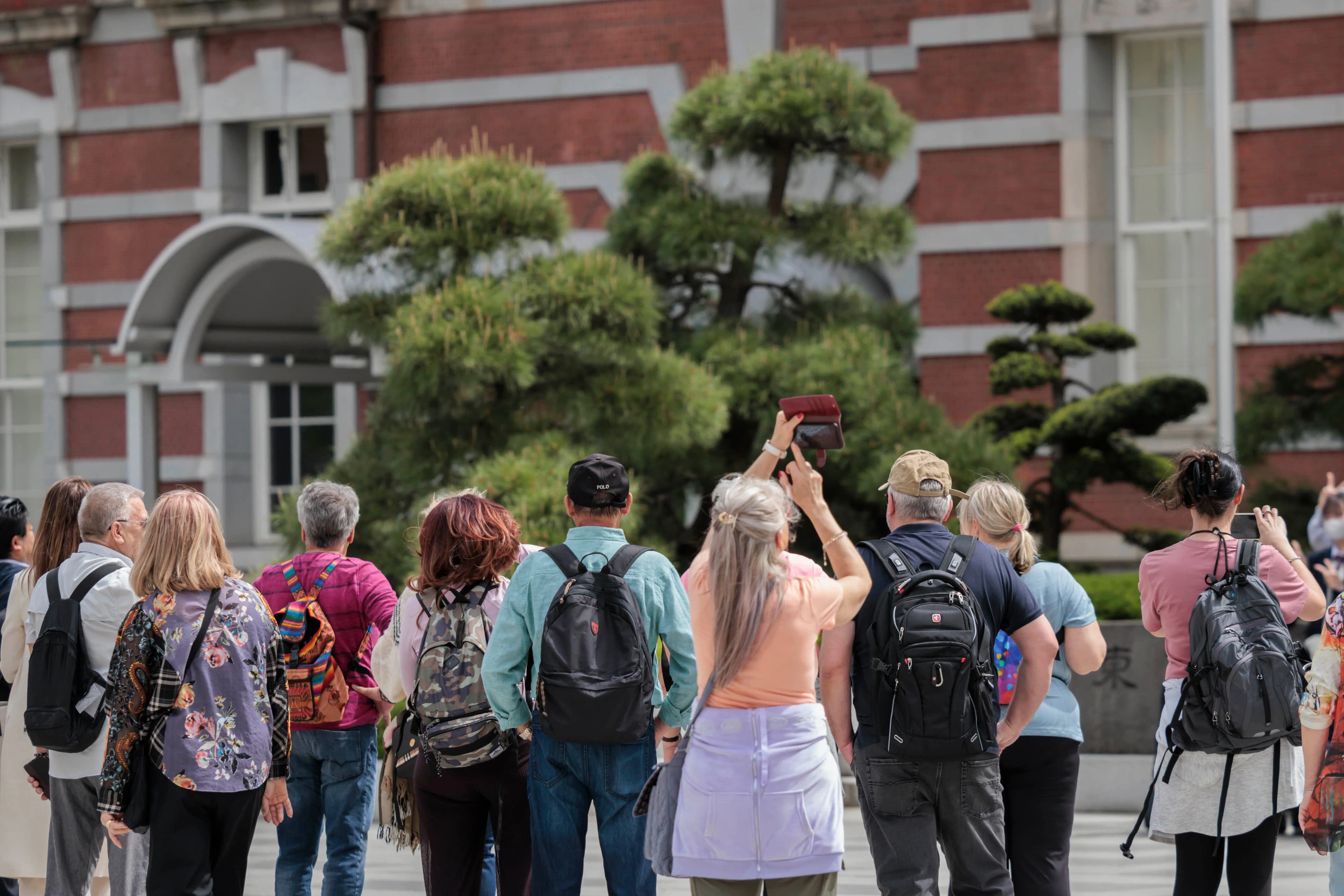 A group of foreign tourists with backpacks gathered in front of Tokyo Station's red-brick Marunouchi building, looking up at it while one takes a photo.